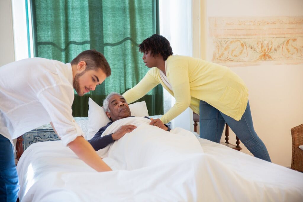 pexels-photo-7551683 Caregivers supporting an elderly man lying in bed, providing assistance and comfort.